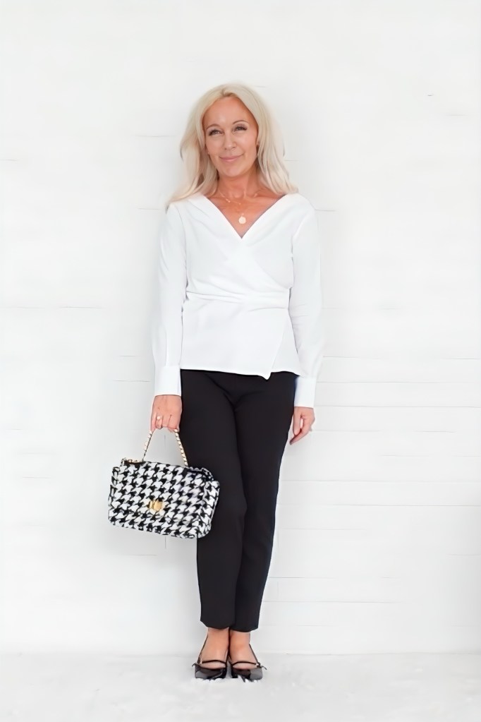 A woman with long, light-colored hair stands in front of a white wall, wearing a white wrap top and black trousers, holding a black and white houndstooth handbag.