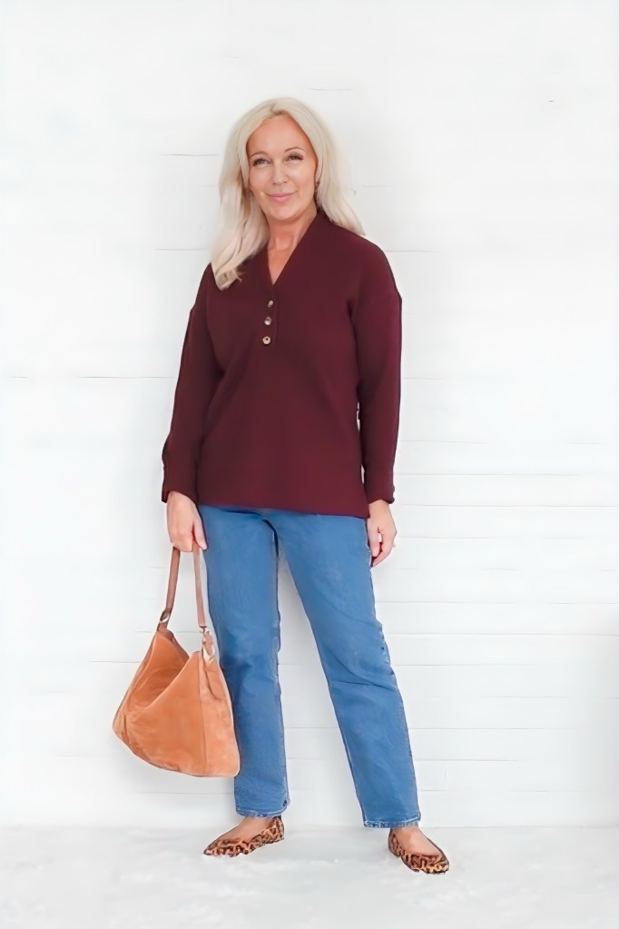 A woman smiling while wearing a burgundy button-up top with three buttons, paired with blue jeans and leopard print flats. She holds a tan bag and stands against a white wooden background.