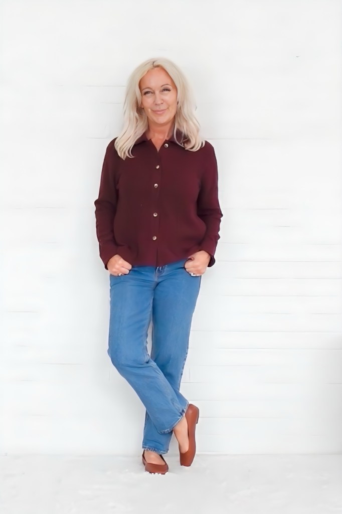 A woman with long blonde hair stands confidently against a white wall, wearing a burgundy button-up shirt and blue jeans, with a relaxed expression.