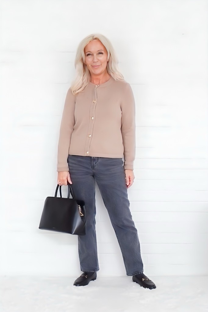 A woman with long blonde hair stands against a white wall, wearing a beige cardigan and gray jeans, holding a black handbag.