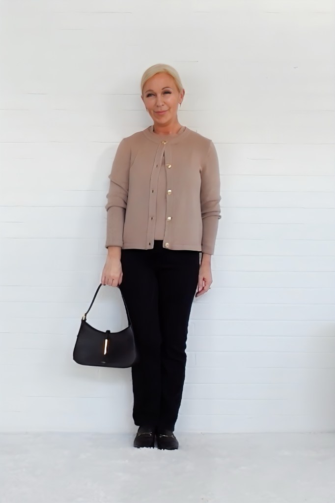 A woman in a beige cardigan and black pants stands against a white wall, holding a black handbag and smiling.