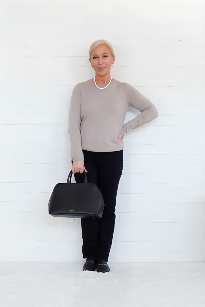 A woman standing against a white background, wearing a beige long-sleeve top and black pants, holding a black handbag.