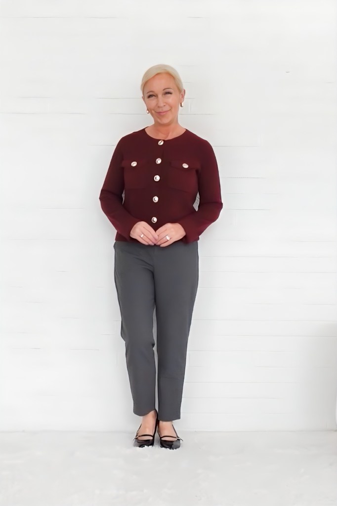 A woman stands against a white background, wearing a maroon cardigan with large buttons and gray pants, styled elegantly with black flats.
