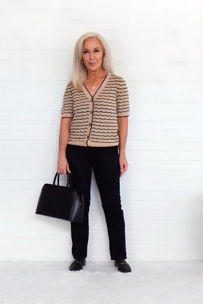 A woman stands against a white wall, wearing a beige patterned cardigan and black pants, holding a black handbag.