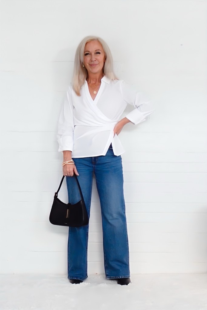 A woman with long hair wearing a white wrap blouse and wide leg jeans, holding a black handbag with a confident smile against a plain white background.