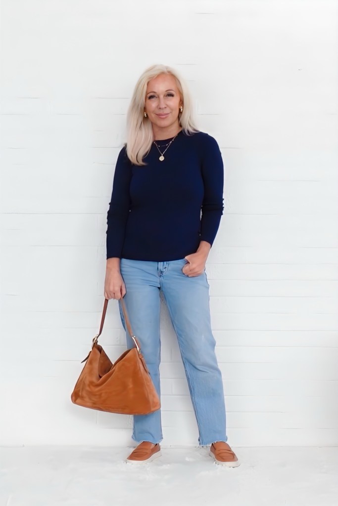 A woman with long blonde hair wearing a navy sweater and light blue jeans holds a brown bag while standing against a white wall.
