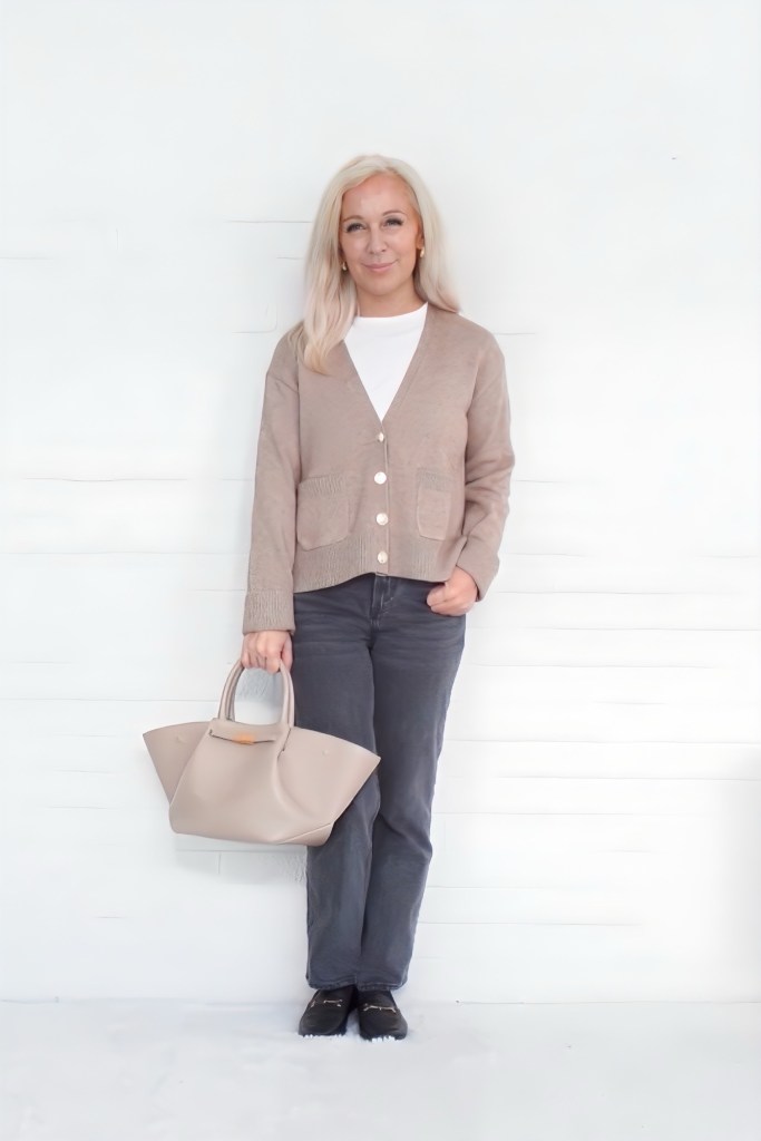 A woman stands in front of a white background wearing a beige cardigan, white top, and dark gray jeans, holding a light beige handbag.