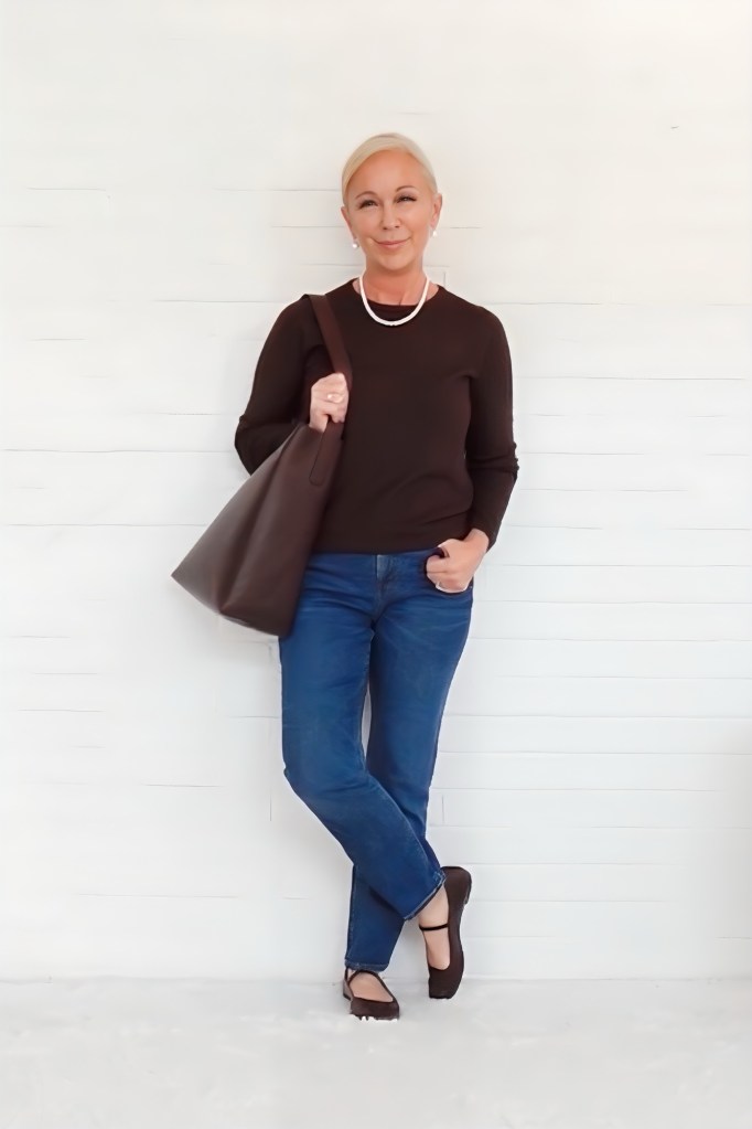A confident woman in a casual outfit, featuring a dark sweater, blue jeans, and a large brown tote bag, stands against a white wooden backdrop.