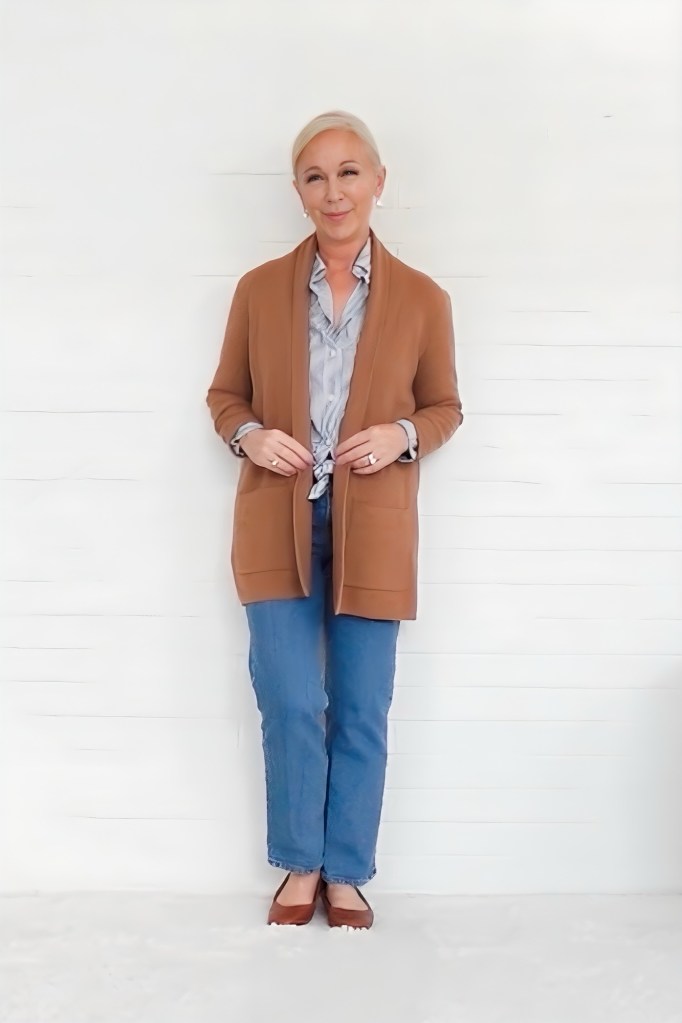 A smiling woman stands confidently in front of a white background, wearing a cozy brown cardigan over a light blue shirt, paired with blue jeans and brown flats.