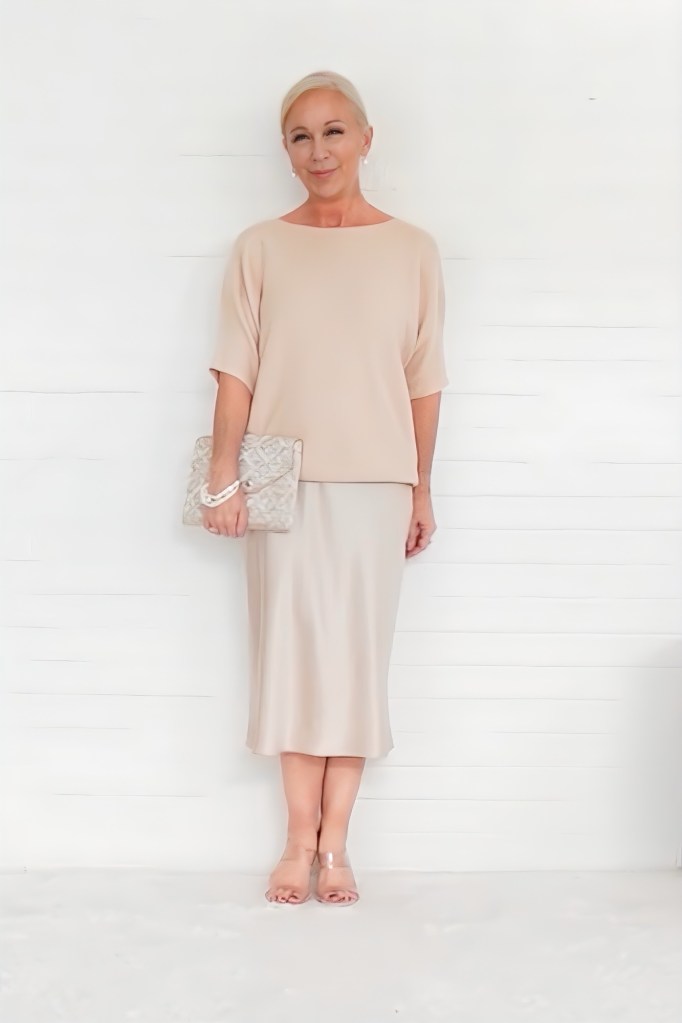 A woman in a neutral-colored outfit stands elegantly against a white background, holding a decorative clutch. Her hairstyle is neat and polished, adding sophistication to her look.