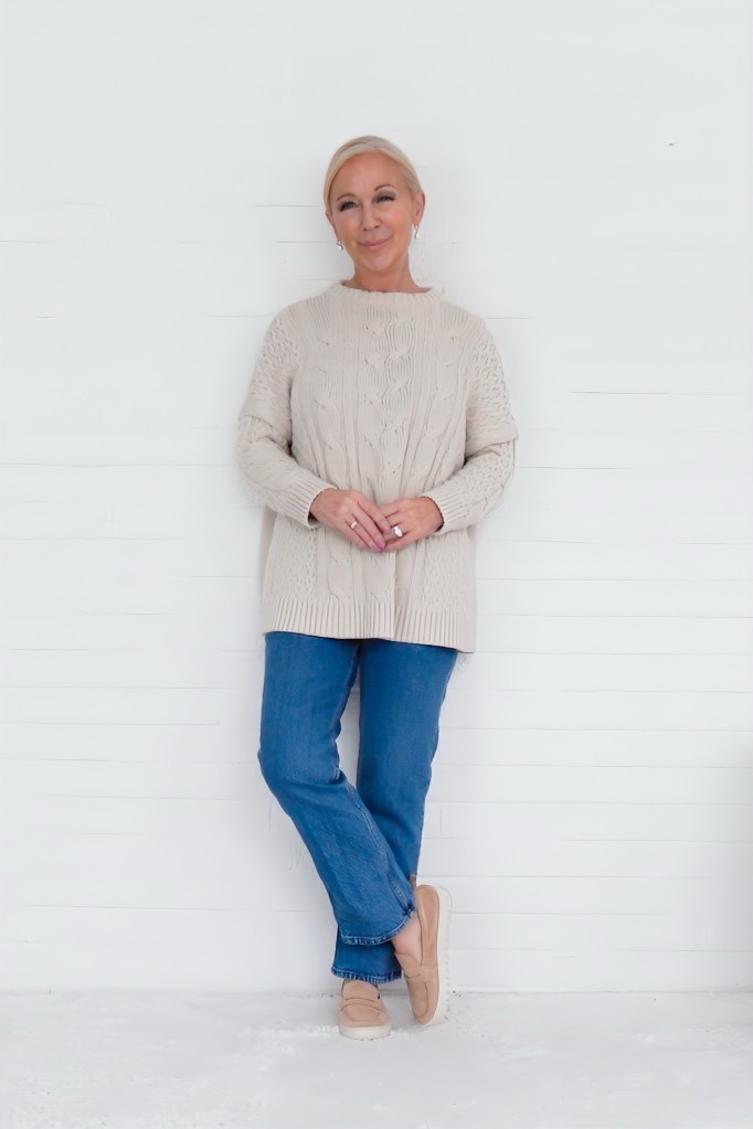 A woman standing in a cozy, chunky cable-knit sweater paired with blue jeans and comfortable shoes, smiling against a white background.