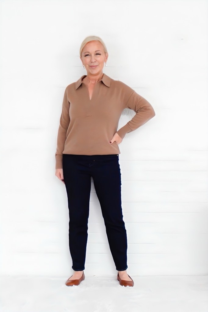A woman standing confidently against a white background, wearing a brown long-sleeve top, dark jeans, and brown flats, with her left hand on her hip and a smile on her face.