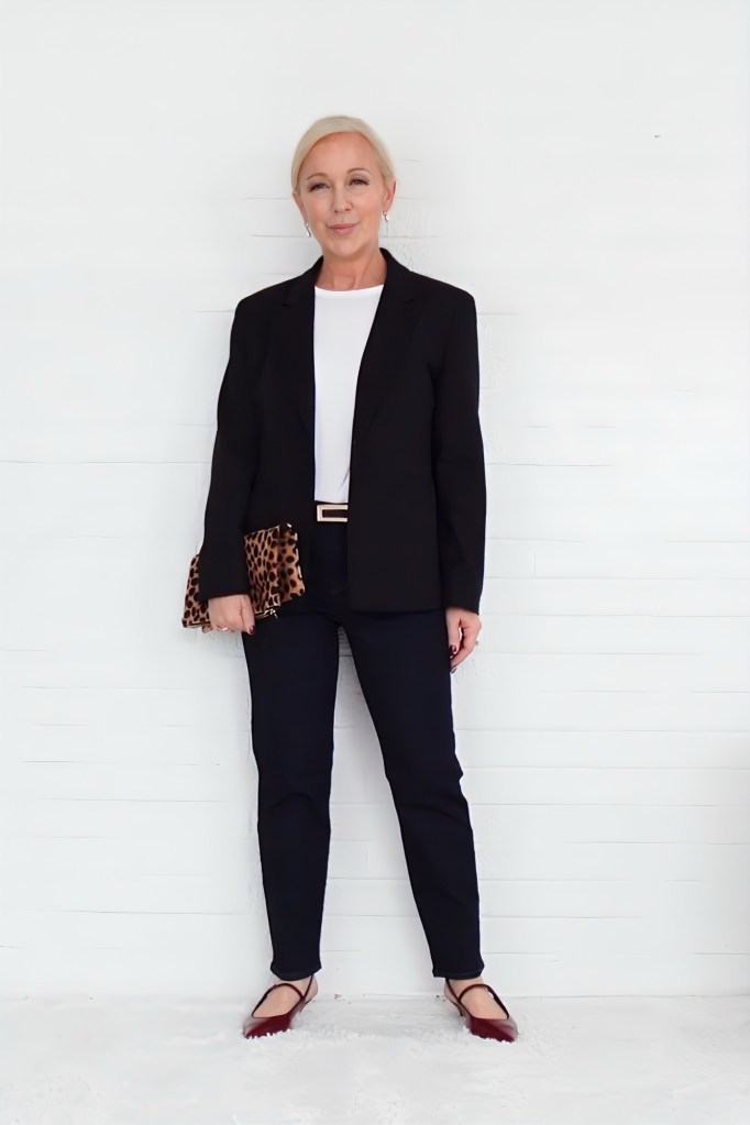 A woman wearing a black blazer over a white top, dark jeans, and burgundy shoes, holding a leopard print clutch while standing against a white background.