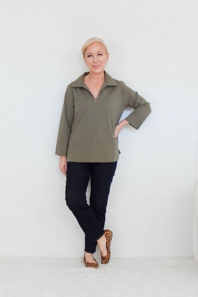 A woman with blonde hair smiles while standing against a white wall, wearing an olive-green blouse and dark pants, paired with leopard-print flats.