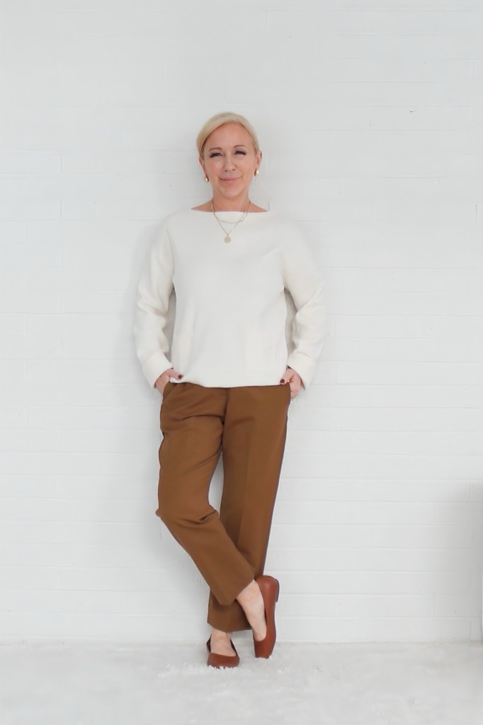 A woman stands against a white brick wall, wearing a cream-colored sweater and brown pants, with a relaxed and confident expression.