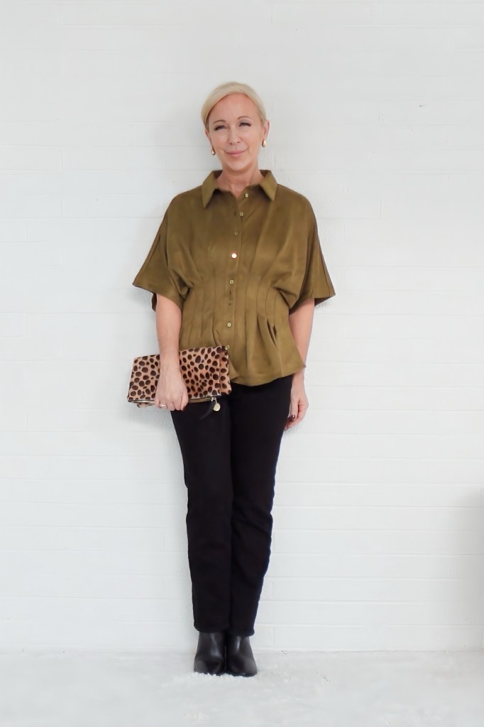 A woman with shoulder-length blonde hair stands against a plain white wall, wearing an olive-green blouse and black pants. She holds a leopard-print clutch and has a confident smile.