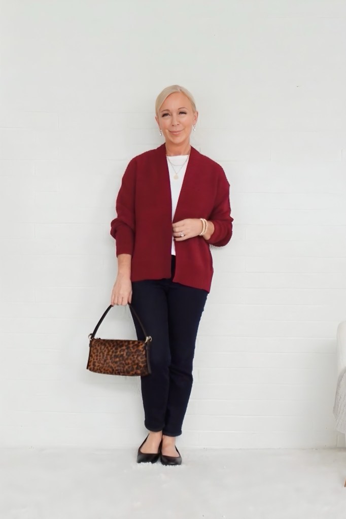 A woman standing against a white wall, wearing a burgundy cardigan over a white top, paired with dark jeans and black flats. She is holding a leopard print handbag and smiling.