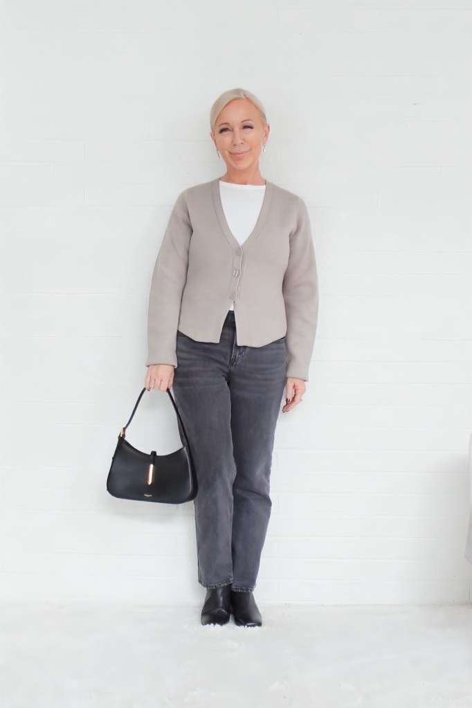 A person wearing a light gray cardigan over a white top, paired with gray jeans and black ankle boots, holding a black handbag, standing against a white wall.