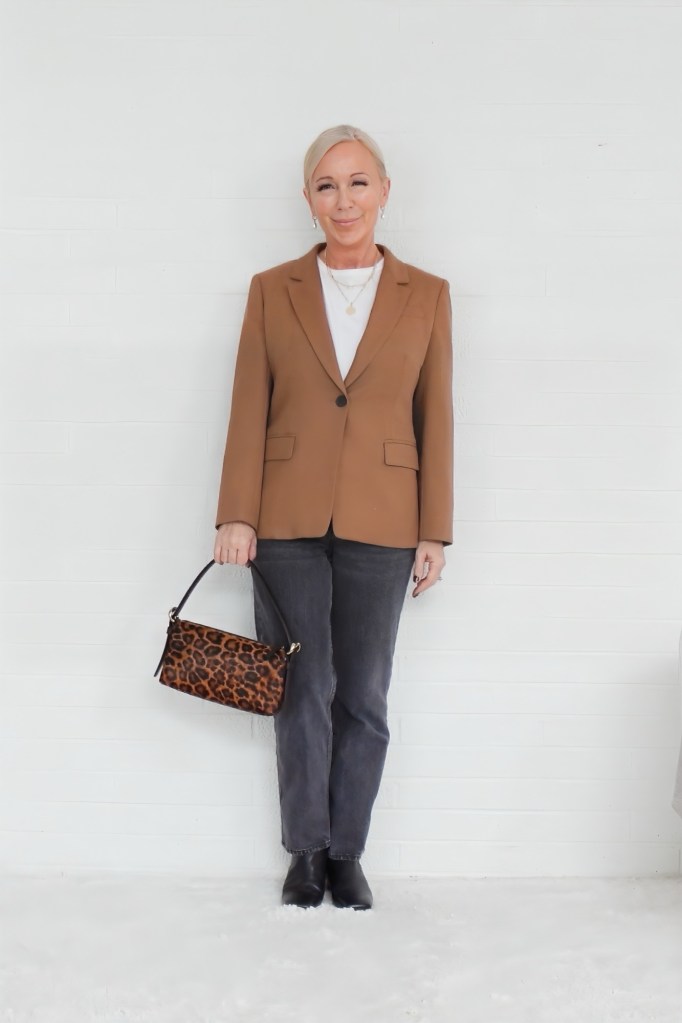 A woman wearing a brown blazer, white top, and gray jeans, holding a leopard print handbag, standing against a white background.