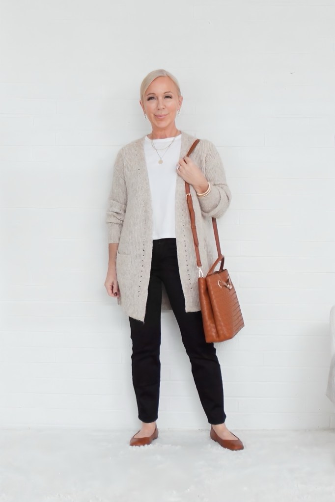 A woman standing against a white wall, wearing a beige cardigan over a white top, black pants, and brown flats, holding a brown handbag.