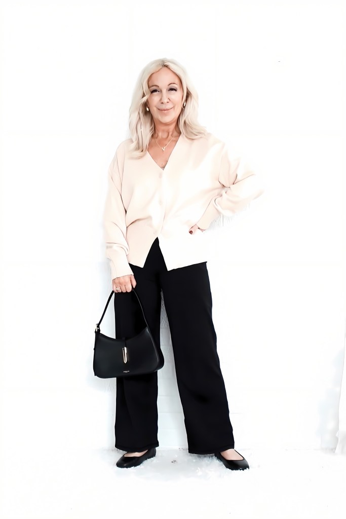 A woman stands confidently in a soft peach cardigan and black wide-leg pants, holding a black handbag. The background is white, creating a clean and minimalist look.