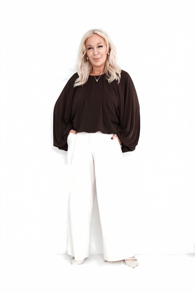 A woman with long, blonde hair stands against a white background, wearing a loose-fitting brown top and white wide-leg pants. She is posing with her hands in the pockets of her trousers, presenting a confident and stylish appearance.