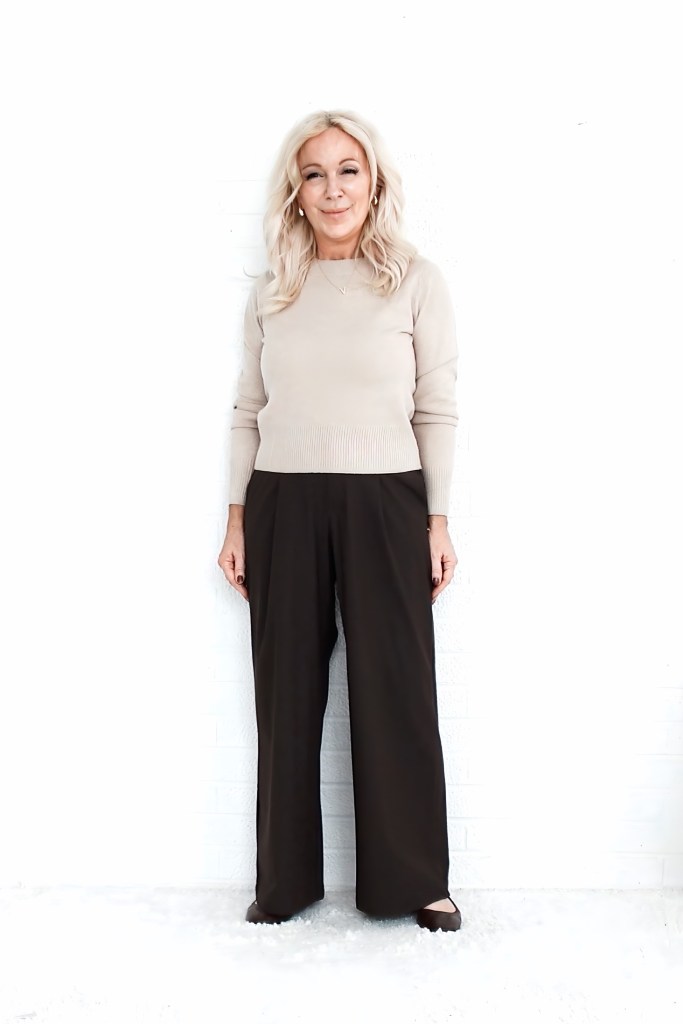 A woman stands smiling against a white background, wearing a light beige sweater and wide-leg brown trousers. She has long, light hair and is accessorized with earrings.