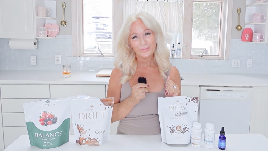 A woman in a kitchen holds a brush with various health supplement packages displayed on the counter, including Bravenly Balance, Drift, Brew, and Calm.