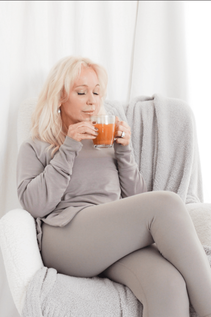 A woman sitting in a cozy chair, holding a glass of tea or beverage, wearing comfortable gray clothing, with a tranquil expression.