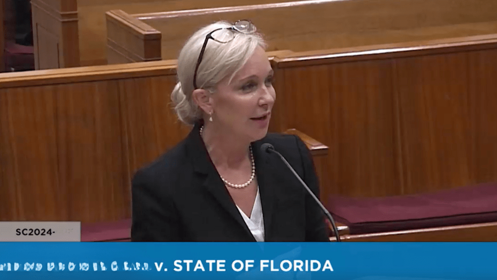 A woman in a professional black suit and pearl necklace speaks at a podium in a courtroom, with wooden paneling in the background.