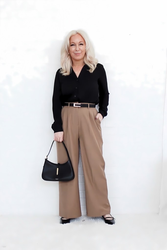 A woman with long blonde hair stands in front of a white wall, wearing a black button-up shirt and tan wide-leg trousers. She holds a black handbag and wears black flats.