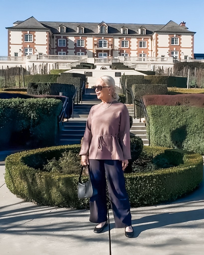 A fashionable woman stands in a manicured garden with a large building in the background. She wears a beige top and navy pants, holding a small black bag.