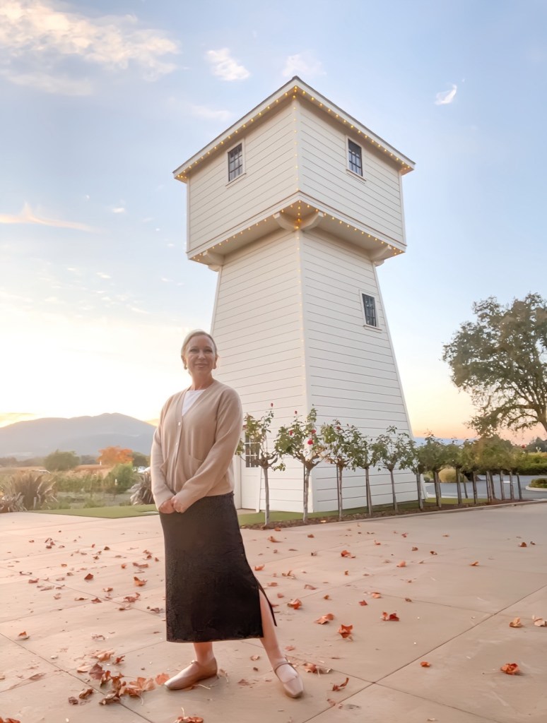 A woman stands smiling beside a white decorative tower adorned with lights, with a backdrop of mountains and a sunset. The ground is covered with fallen leaves.