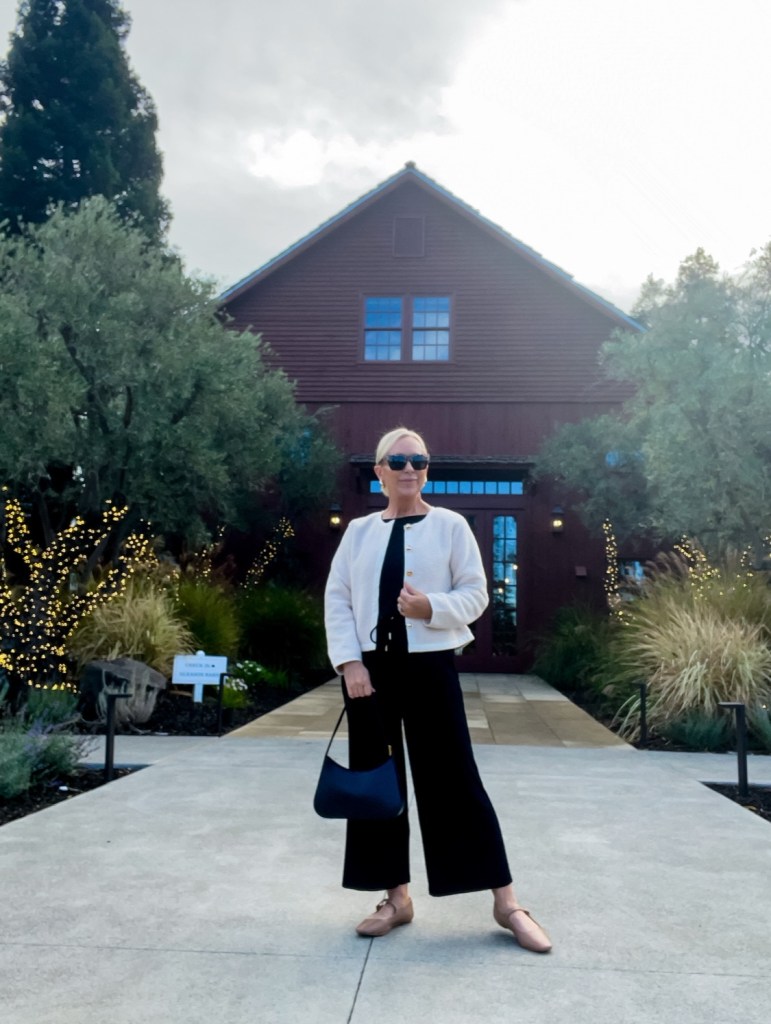 A woman standing in front of a rustic red barn, wearing sunglasses, a white jacket, and black wide-leg pants, holding a black bag, with trees and fairy lights in the background.