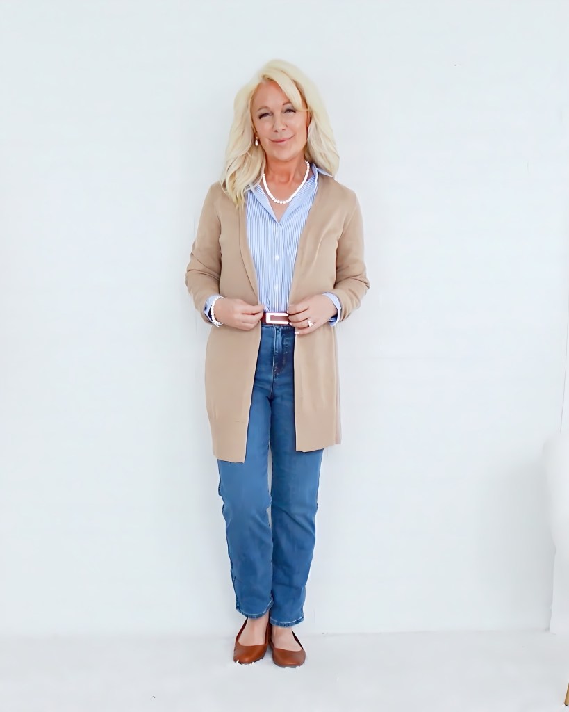 A woman standing in front of a plain white background, wearing a beige cardigan over a blue and white striped shirt, with blue jeans and brown shoes, smiling gently.