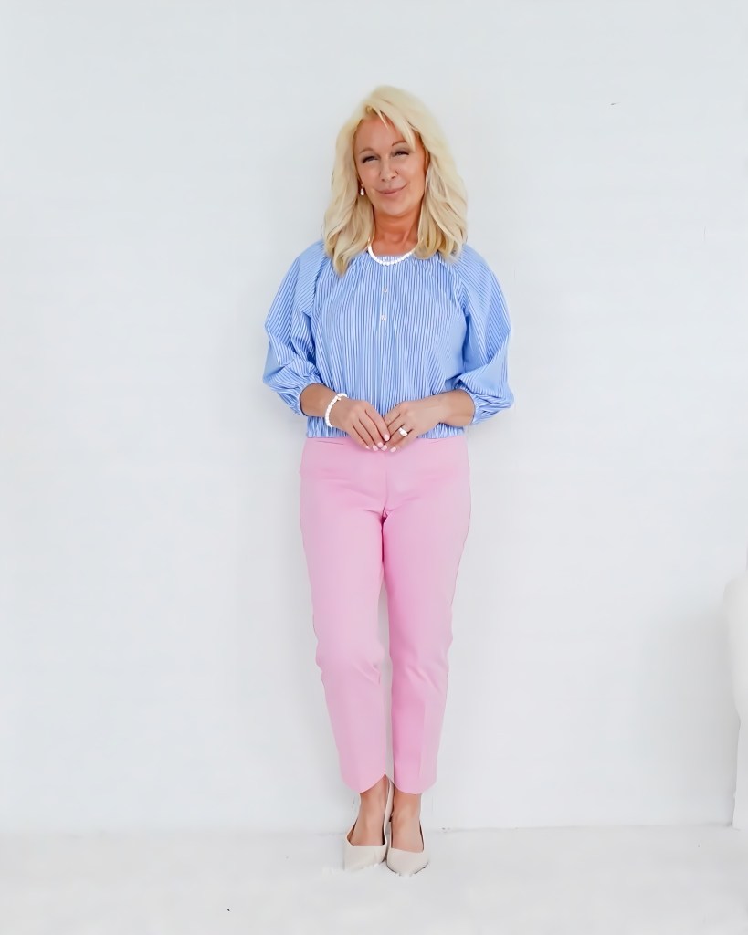 A woman standing against a white background, wearing a blue-and-white striped blouse and pink pants, in a confident pose and with a friendly expression.