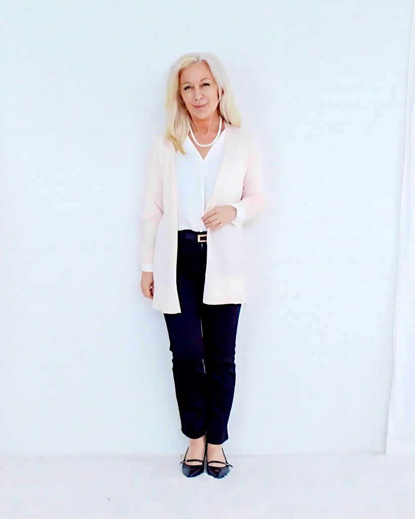 A woman with long, light blonde hair wearing a white blouse, light pink cardigan, and dark pants, stands against a white background, smiling gently.