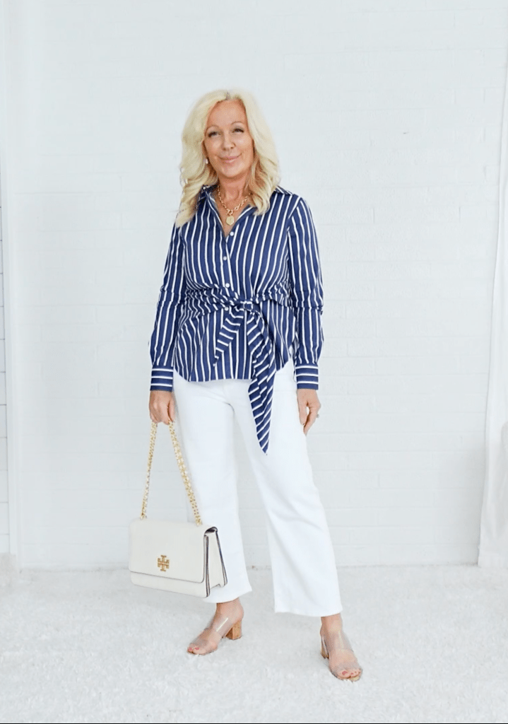 Woman standing against a white background, wearing a striped navy blue shirt tied at the waist and white pants, holding a white handbag with a gold chain strap.