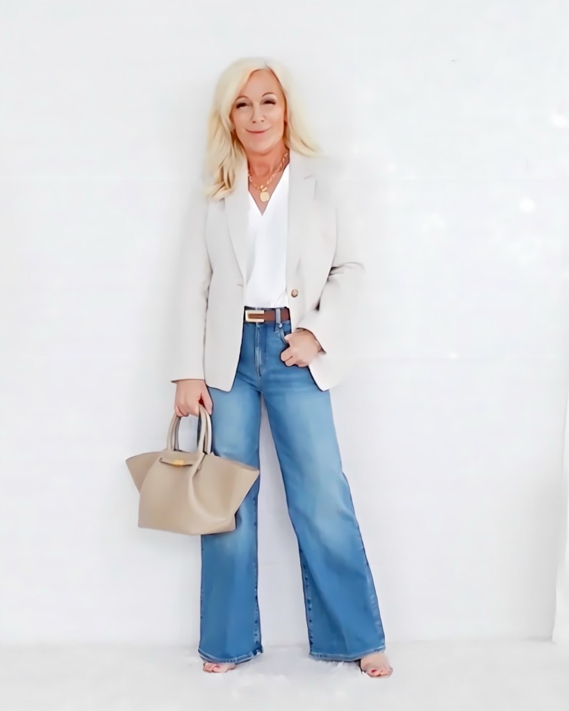 A woman stands smiling against a white background, wearing a light gray blazer over a white blouse, paired with wide-leg blue jeans. She holds a beige handbag in one hand.