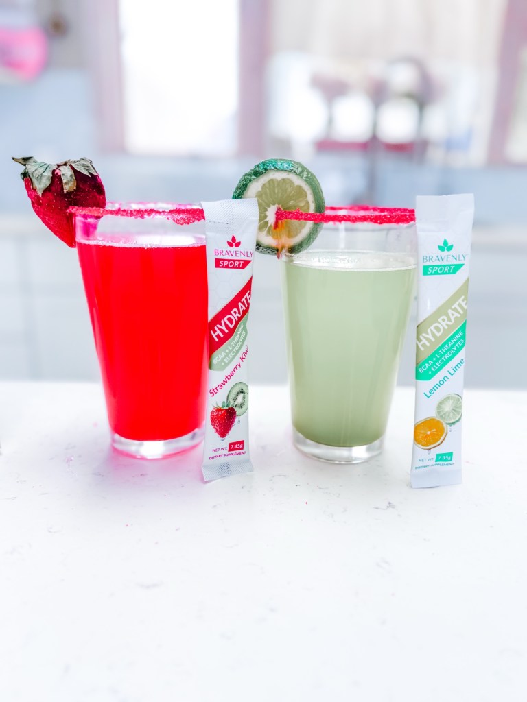 Two colorful drinks in clear glasses: a red drink garnished with a strawberry and a green drink with a lime slice, accompanied by packets of hydration supplements labeled 'Strawberry Kiwi' and 'Lemon Lime'.