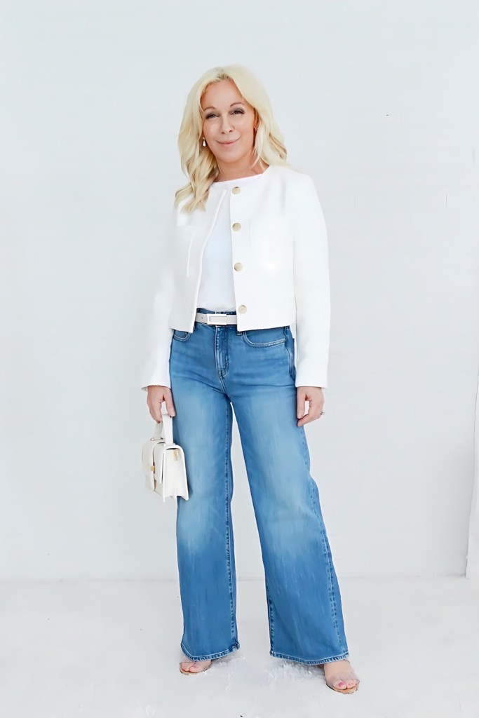 A woman stands confidently in a stylish outfit featuring a white cropped jacket with gold buttons, a light top, and flared blue jeans. She holds a small white handbag and poses against a light background.
