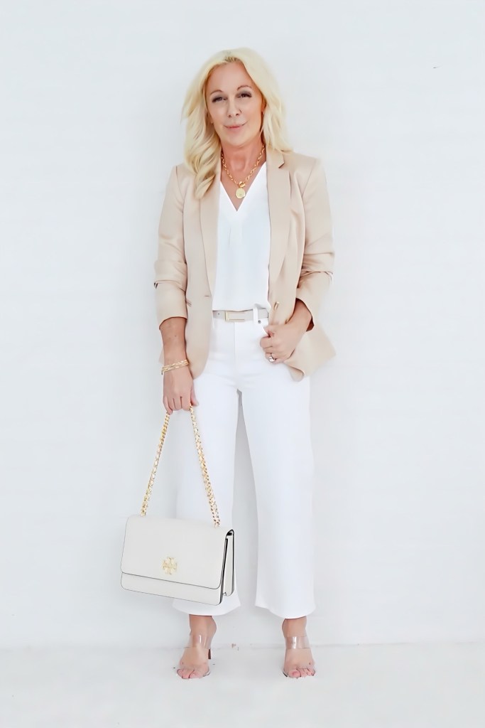 A woman stands against a plain white background, wearing a beige blazer over a white top and white wide-leg pants. She carries a light-colored handbag with a gold chain strap and wears clear sandals.