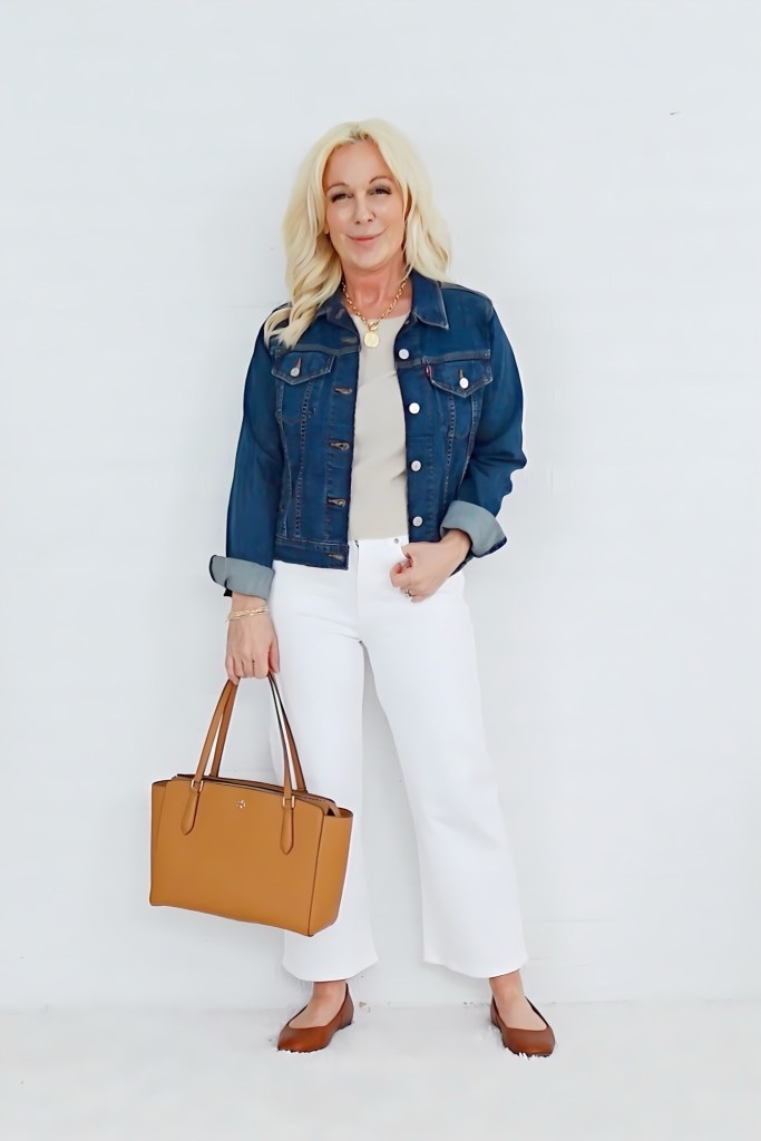 A woman standing against a white background, wearing a denim jacket over a light top, white wide-leg pants, and brown flats. She is holding a tan handbag and smiling.