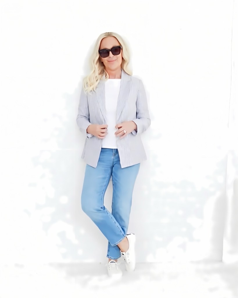 A woman wearing a striped blazer, white top, and light blue jeans stands with one leg crossed against a white wall, smiling and wearing sunglasses.
