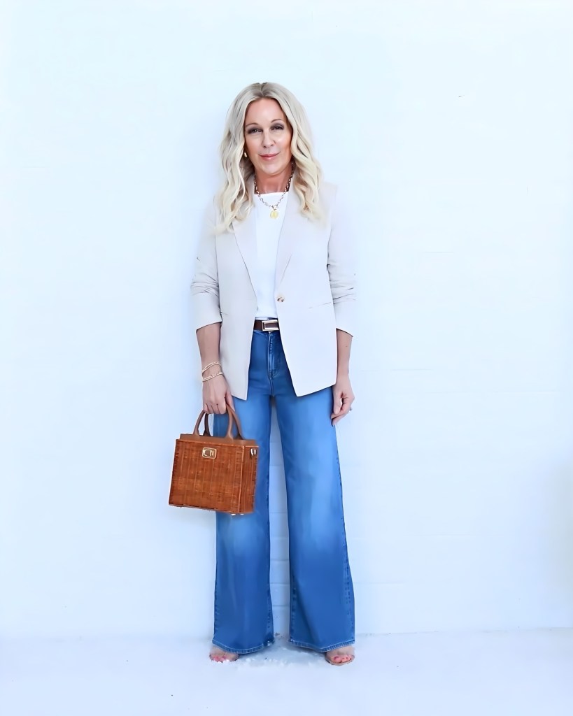 A fashionable woman stands against a white wall, wearing a light-colored blazer, a white top, and wide-leg blue jeans. She holds a small brown handbag and has styled wavy hair.