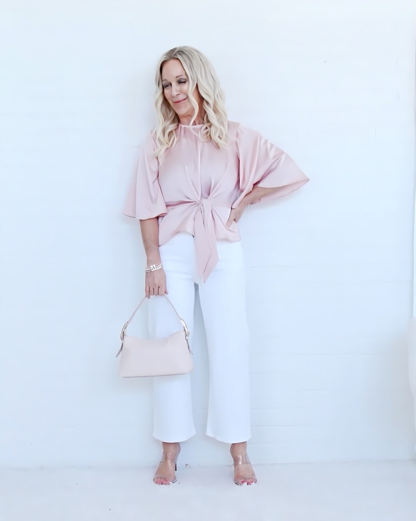 A woman with long blonde hair stands against a white background, wearing a pink blouse tied at the waist and white wide-leg pants. She holds a light beige handbag and has a cheerful expression.