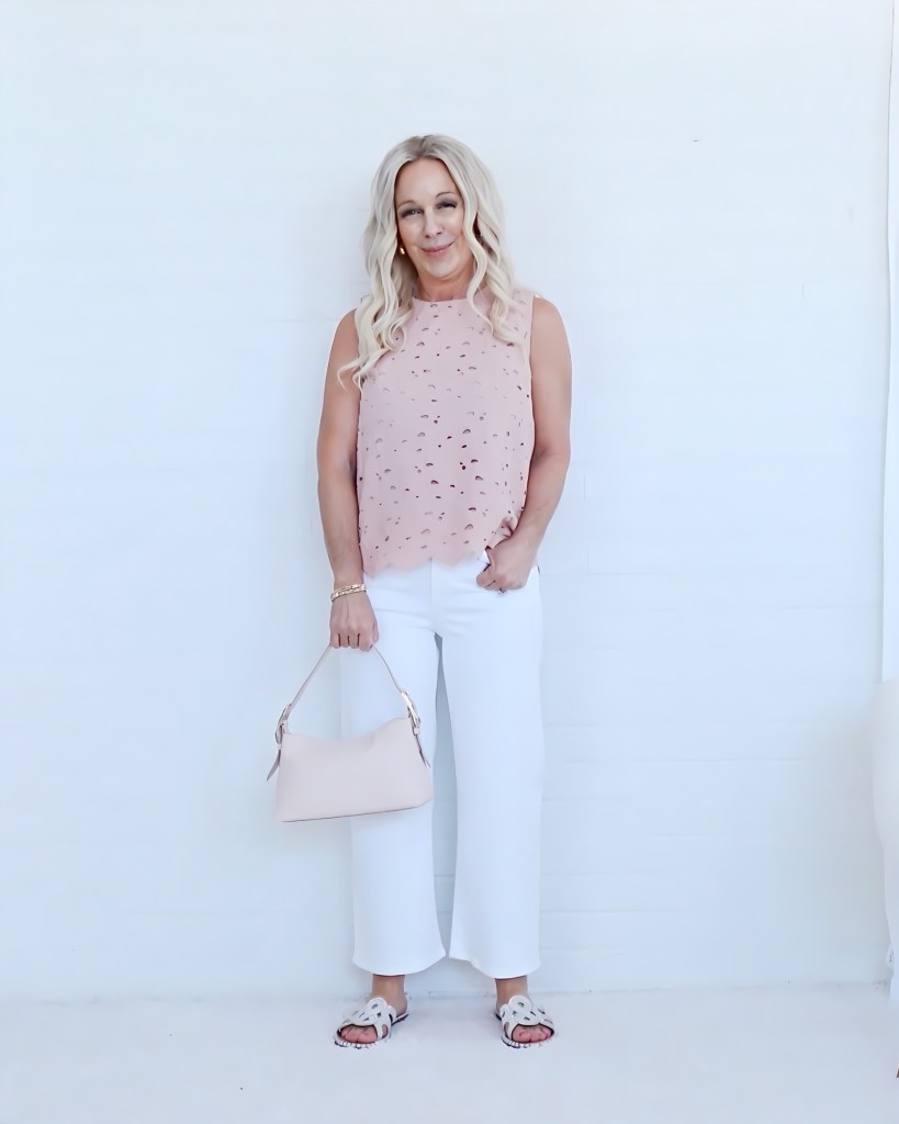 Woman standing against a white wall, wearing a sleeveless pink top, white wide-leg pants, and holding a small nude handbag.