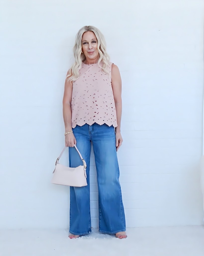 Woman standing against a white wall, wearing a pink sleeveless top with cutout details and blue wide-leg jeans, holding a light pink handbag.
