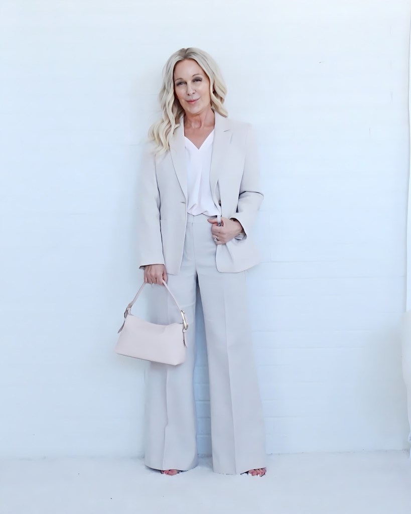 A woman in a light beige suit with wide-leg pants, holding a small matching handbag, standing against a white background.