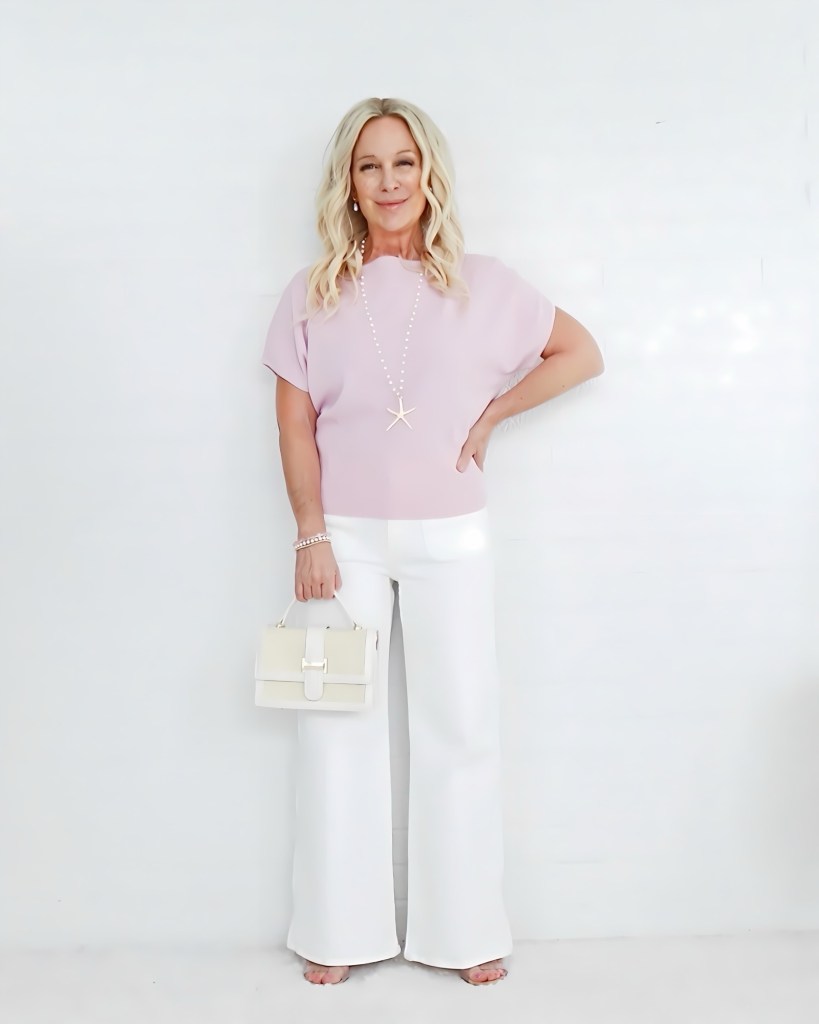 A woman stands in front of a white background, wearing a light pink top and wide-leg white pants. She has long, wavy blonde hair and is holding a small beige handbag. She's accessorized with a starfish necklace and a bracelet.